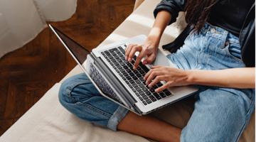 A young person sitting cross-legged on a couch and typing on their laptop