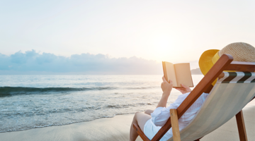 Image of a person on the beach, sitting in a chair reading a book.