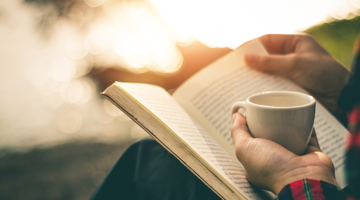 A person sitting outside holding a mug in one hand and a book on their knee.