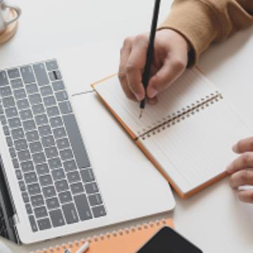 A person at the desk writing notes with the laptop opened